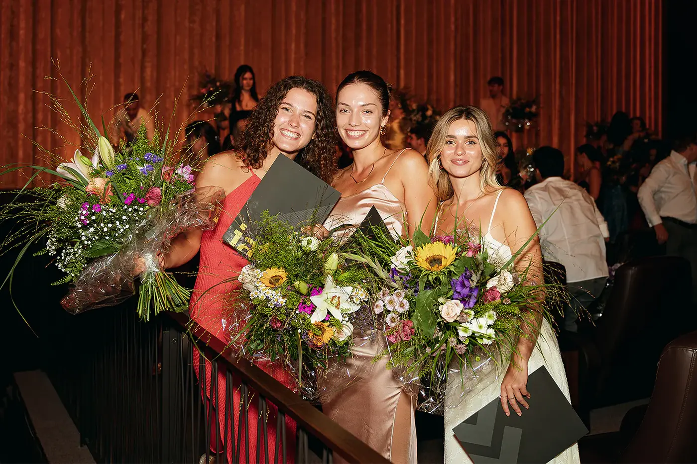 Drei Absolventinnen mit Blumensträußen und Zeugnismappen, IAF Abschlussfeier 2024 in Köln